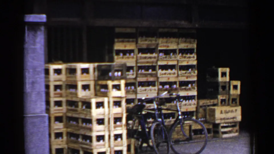 1953 Japan 8mm frame with stacked wooden crates of bottles, a bicycle in front, a stone pillar, and additional crates along the wall.