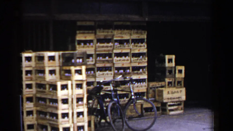 1953 Japan 8mm frame showing a bicycle parked before stacked wooden bottle crates, with a dark storage opening and pavement in view.