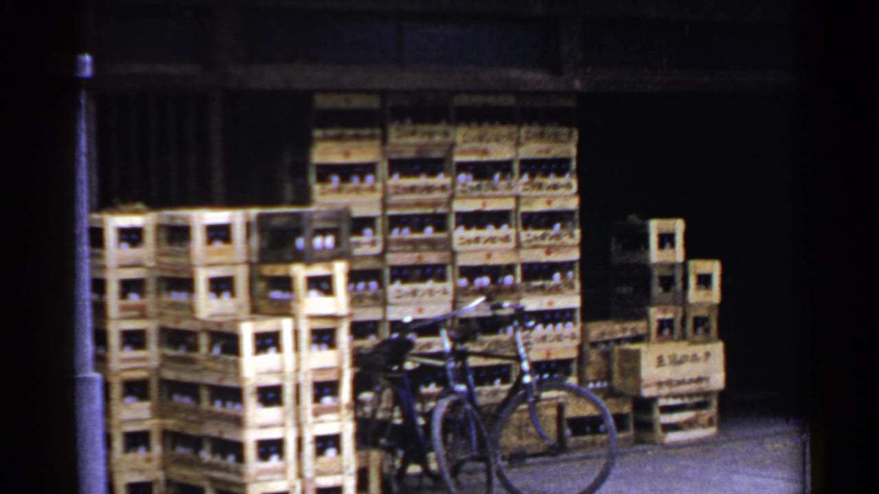 Stacked wooden crates filled with bottles beside a bicycle at a shadowed storefront or warehouse.