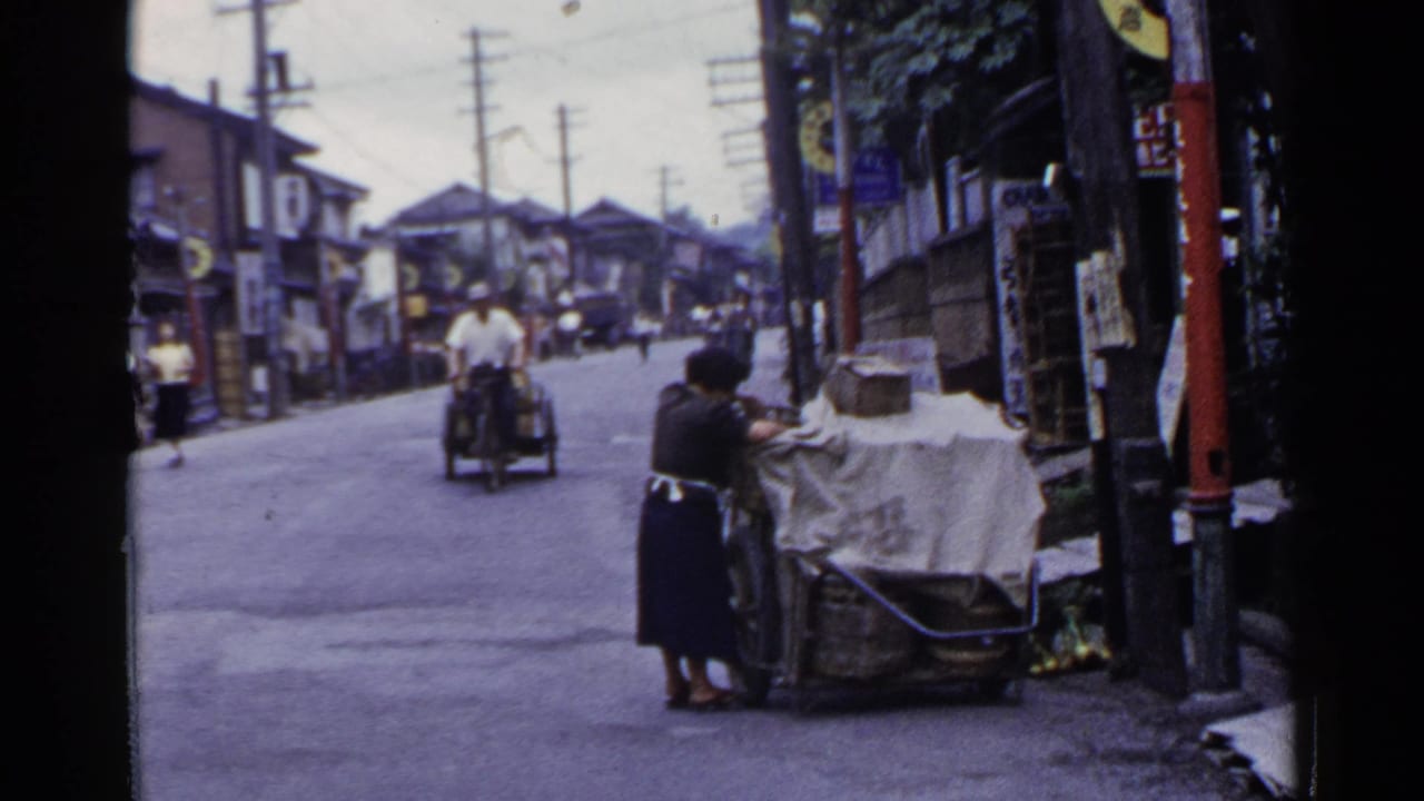 1953 Japan street view with a woman tending a covered cart while a motorized cycle-cart approaches between wooden buildings.