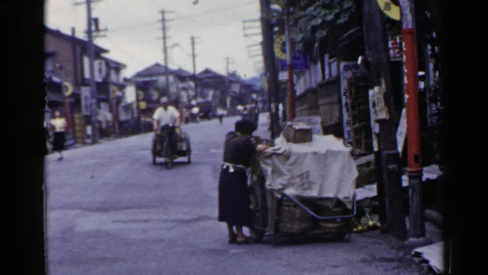 1953 Japan street view with a woman tending a covered cart while a motorized cycle-cart approaches between wooden buildings.