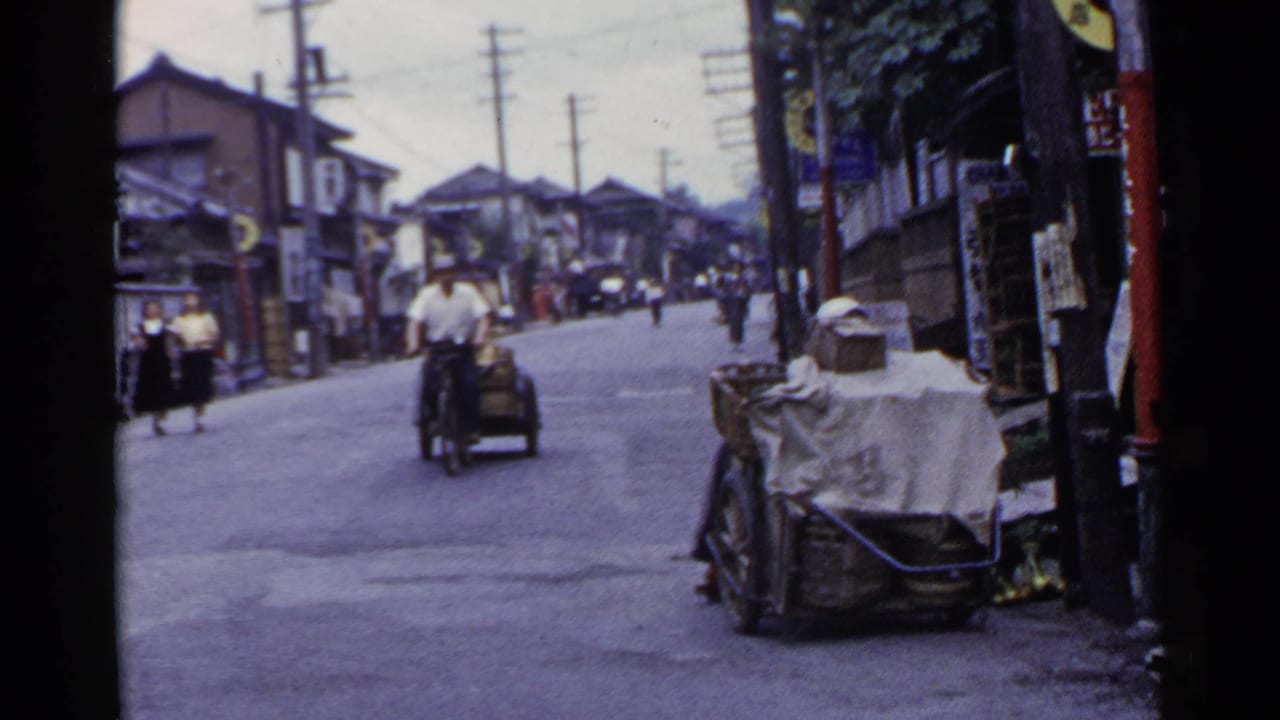 A rider on a small motorized cycle-cart passes along a 1953 Japanese street with pedestrians and signs in view.