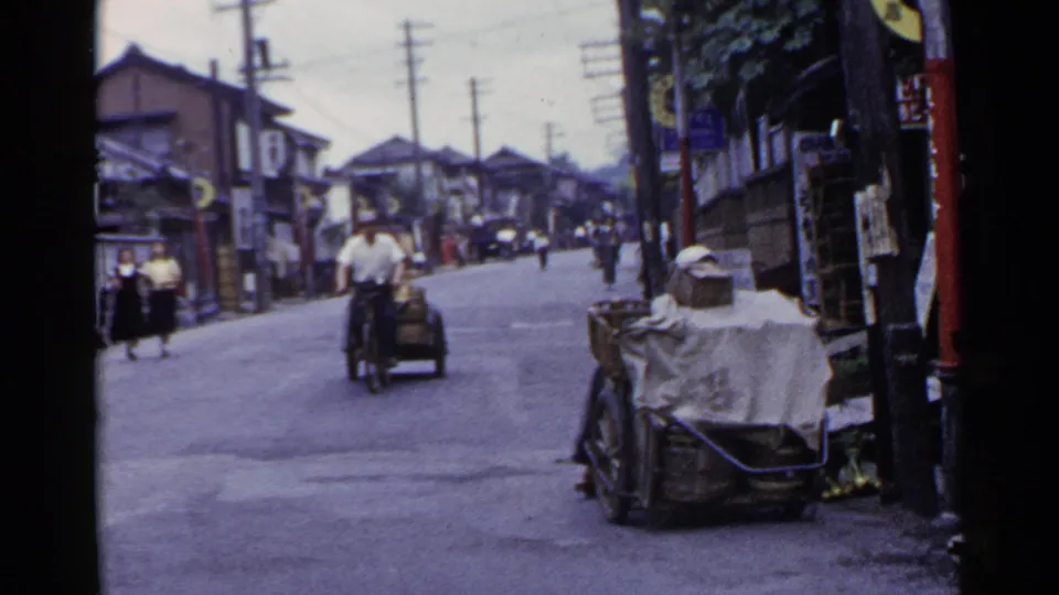 A rider on a small motorized cycle-cart passes along a 1953 Japanese street with pedestrians and signs in view.