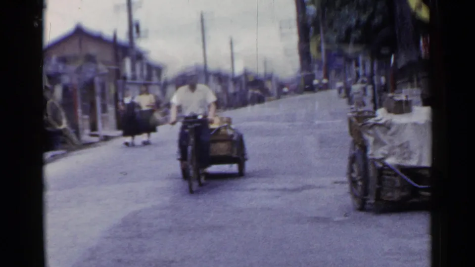 Soft 8mm frame of a cycle-cart rider approaching on a Japanese town road as pedestrians continue in the background.