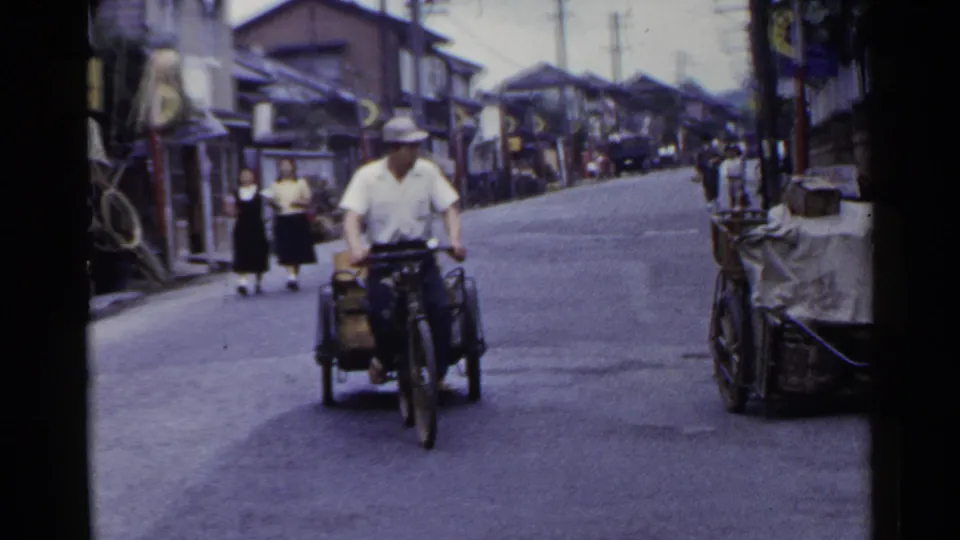 A man rides a motorized cycle-cart near the camera on a 1953 Japan street, with two uniformed schoolgirls walking in the distance.