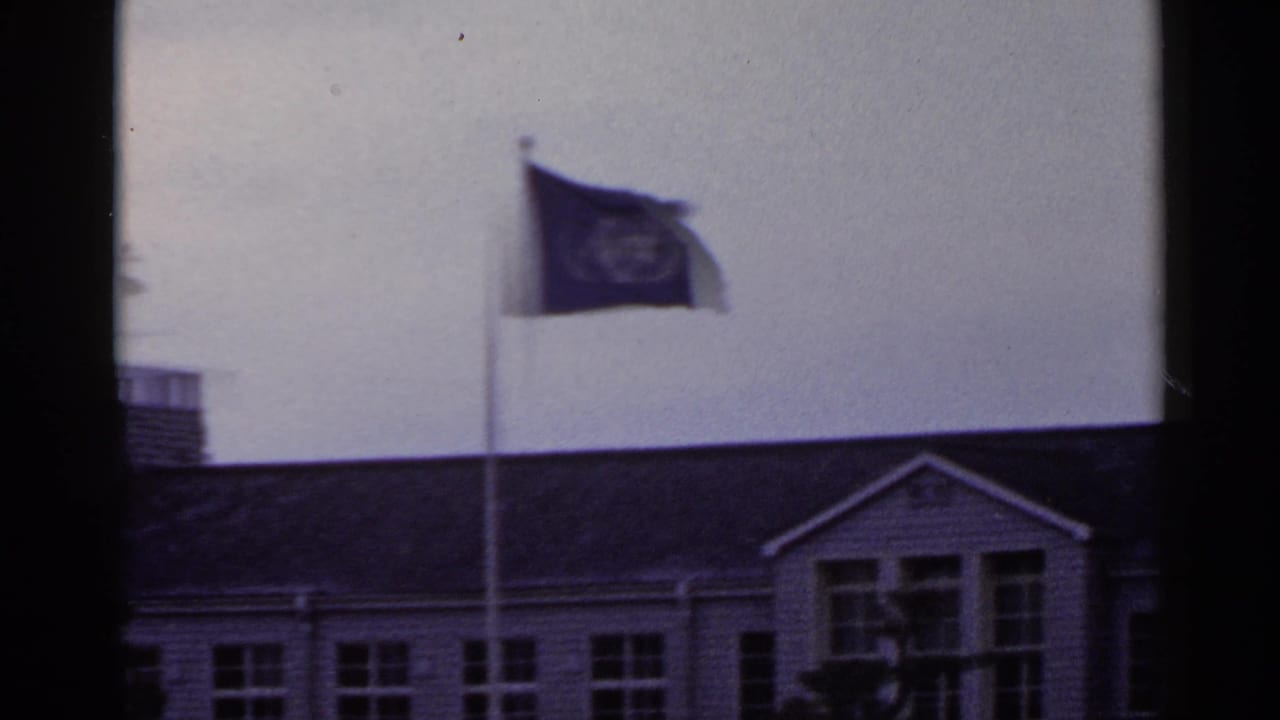 1953 Japan 8mm frame showing a United Nations flag on a pole above a building with windows and a dark roofline.