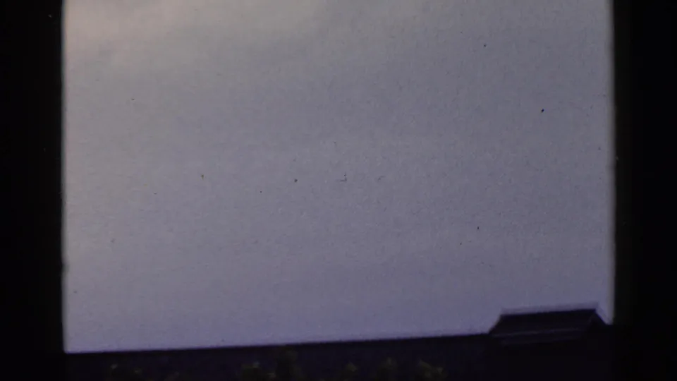 Soft 8mm frame from the pan showing pale sky, film grain, and a small dark roof edge at lower right.