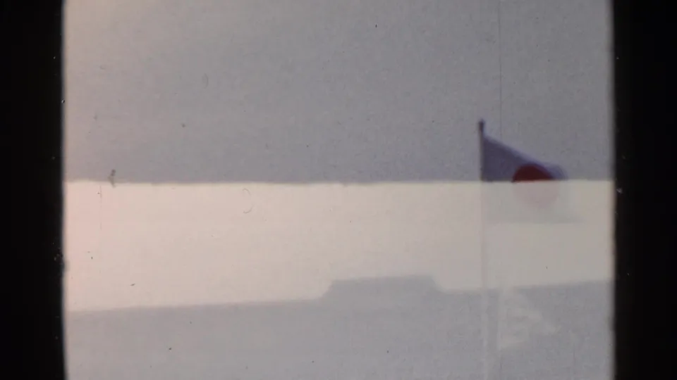 1953 Japan 8mm frame showing a Japanese flag on a flagpole, with a pale sky and distant building roof below.