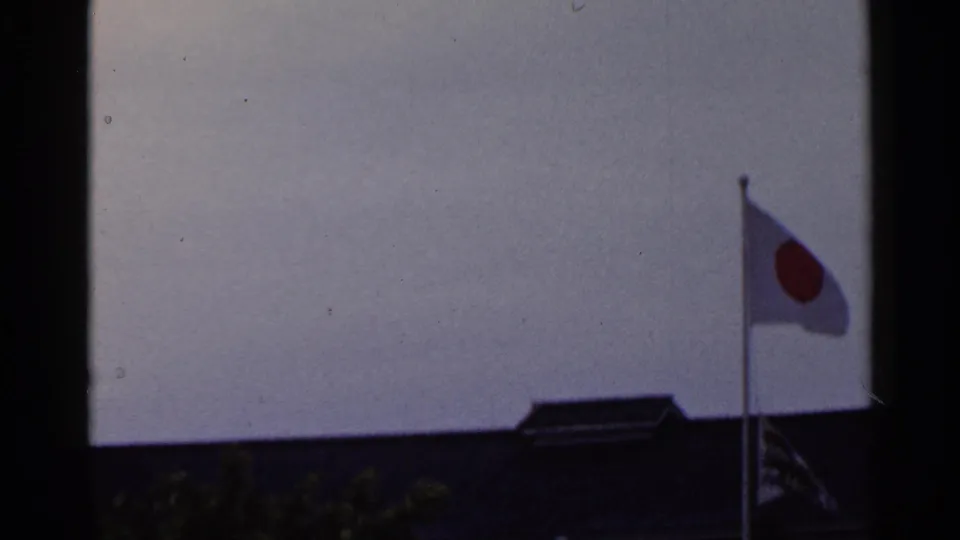 1953 Japan 8mm frame showing the Japanese flag waving above a dark roofline under an overcast sky.