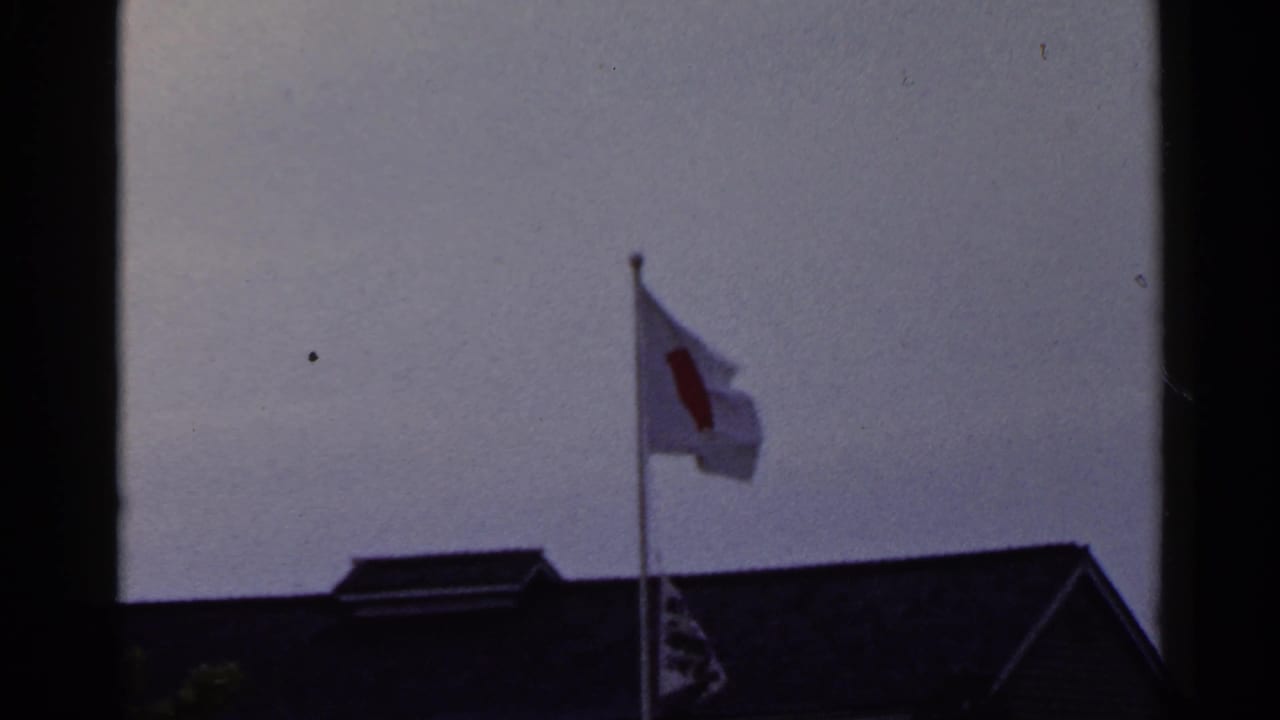 1953 Japan 8mm frame with the Japanese flag bending in the breeze over a dark building roof.