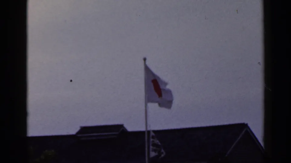 1953 Japan 8mm frame with the Japanese flag bending in the breeze over a dark building roof.