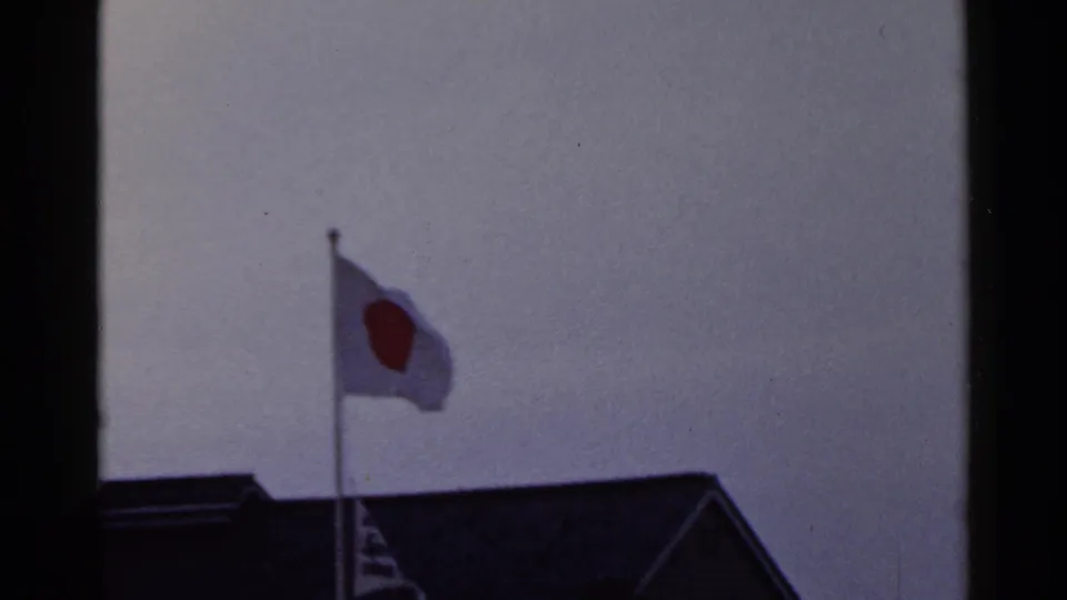 1953 Japan 8mm frame showing the flag's red disc visible as it waves above a dark roofline.