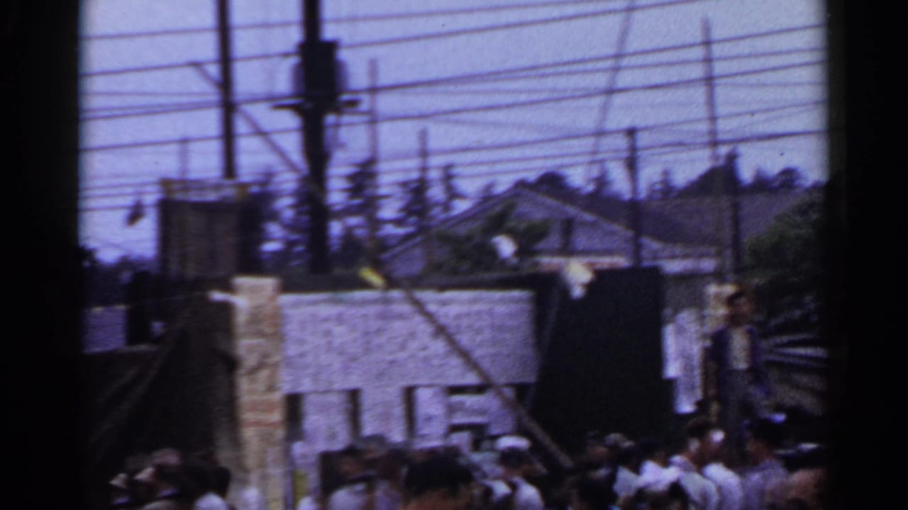 Crowd at a street fair beneath overhead wires, with sailor hats and booth structures visible.