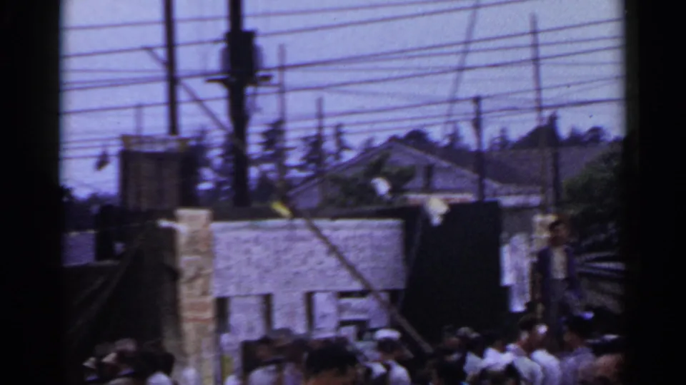 1953 Japan street-fair crowd with sailors in white caps, low buildings, utility wires, and dark 8mm frame edges.