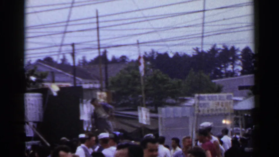 Crowded 1953 Japan fair scene with sailors and civilians moving near stalls beneath many utility wires.