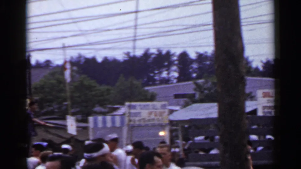 1953 Japan fair view with a painted game sign, crowd, sailor hats, tree trunk, low buildings, and utility lines.