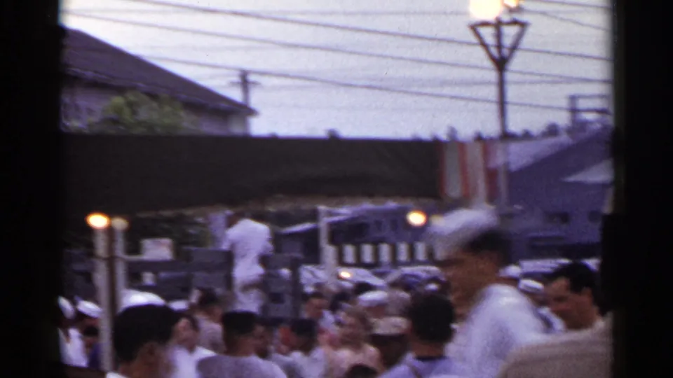 Crowded 1950s Japan street view with pedestrians in light clothing, awnings, overhead wires, and low-rise commercial buildings.