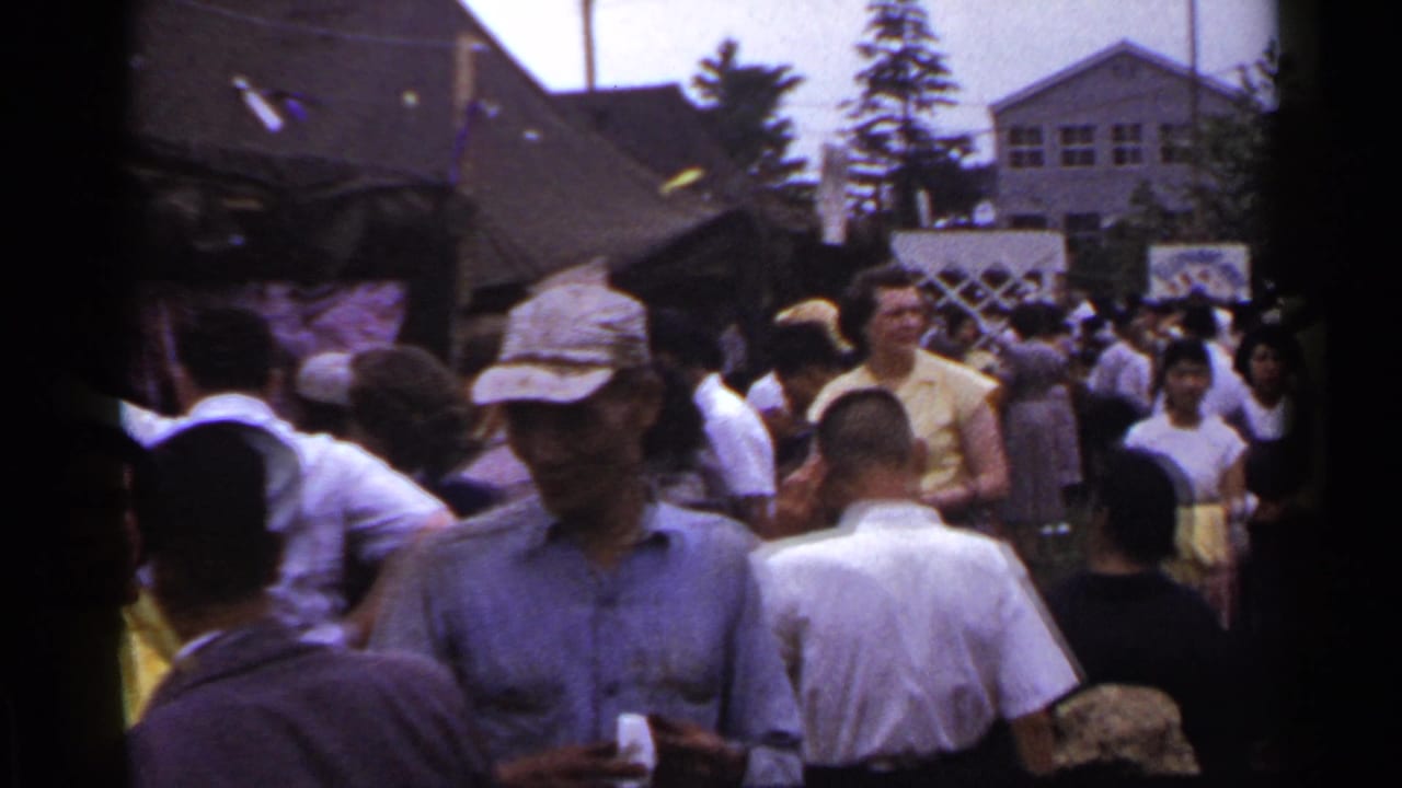 Crowd moving through an outdoor event with tents, buildings, and an American flag in the background.