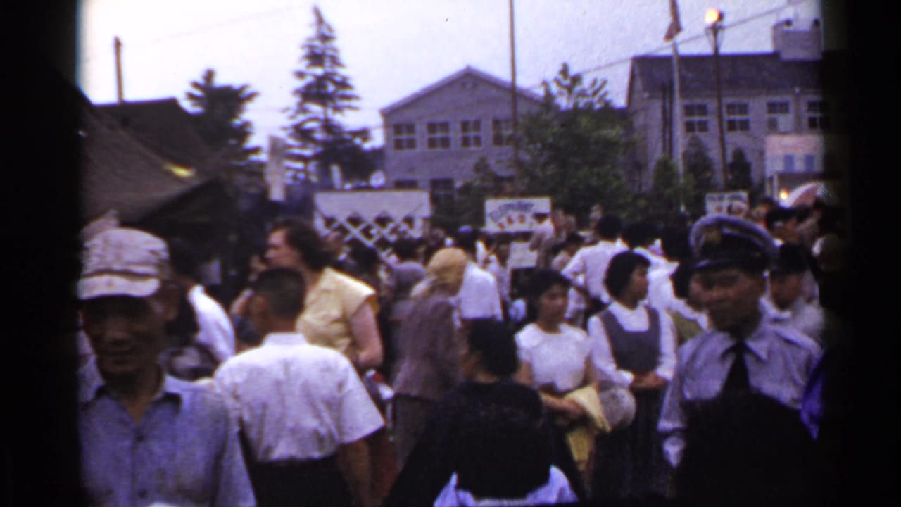 Festival crowd with a uniformed man at right, tents, banners, and buildings in the background.