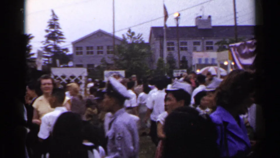 1953 Japan home-movie frame of people walking through an outdoor event with tents, bunting, and large buildings in view.