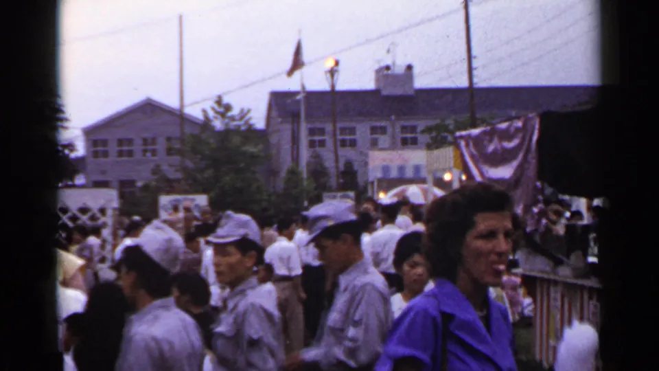 1953 Japan frame showing a crowded outdoor event with men in caps, people near a railing, tents, bunting, and background buildings.