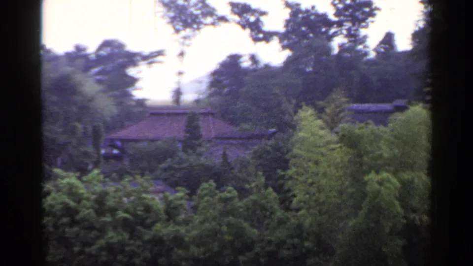 1953 Japan home-movie frame with a tiled-roof house nestled among trees and shrubs; a person is flagged in the clip but not clear here.