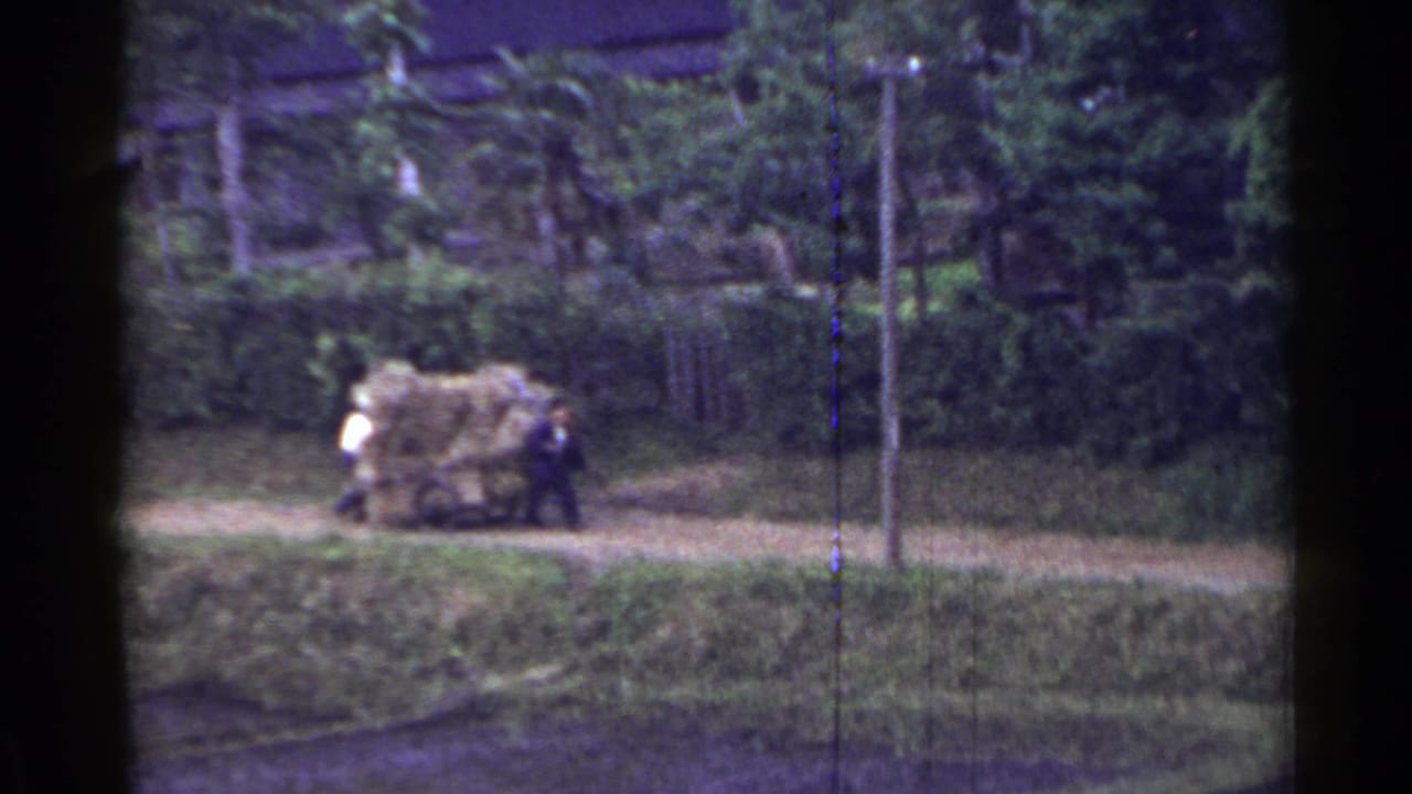 Two men continue pushing a loaded cart past a utility pole on a dirt path.