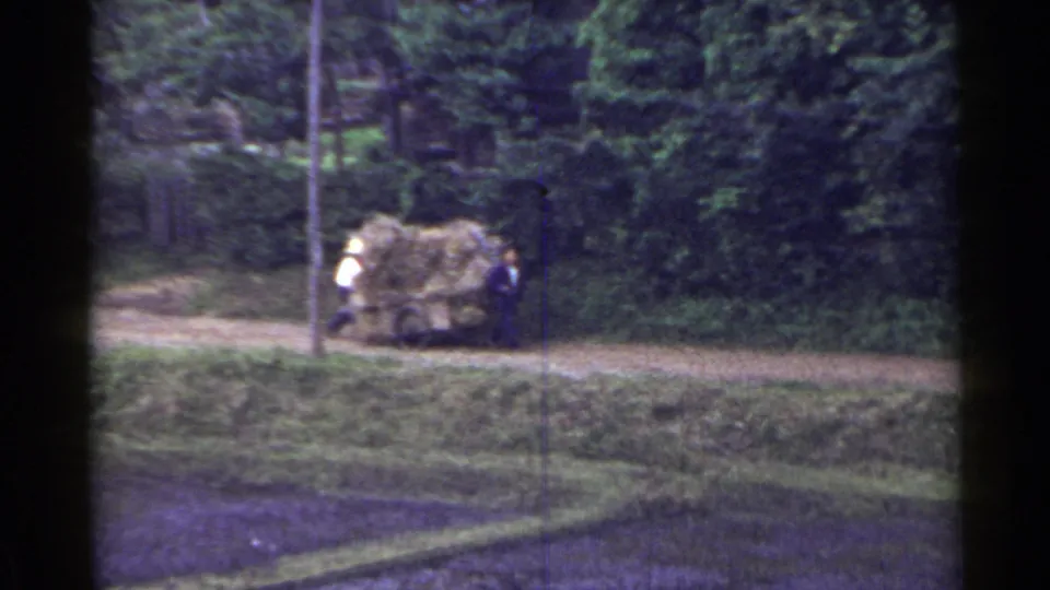 The cart piled high with hay or straw moves along a rural Japanese dirt path, with two men partly visible beside the load.