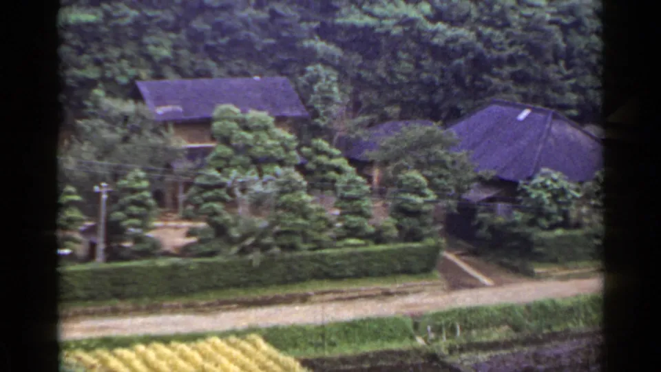 End frame of the 1953 Japan pan showing dark-roofed village buildings, hedges, trees, fields, and a dirt path.