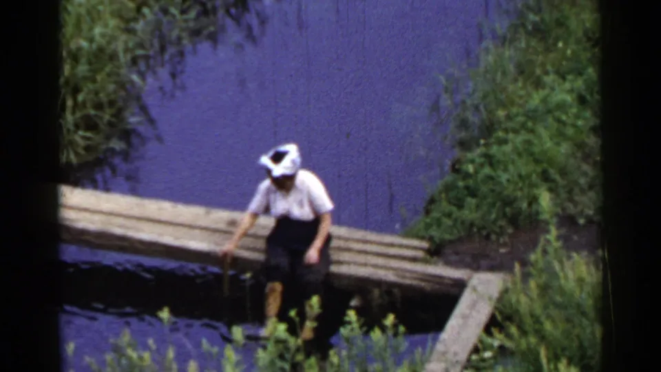 1953 Japan home-movie frame showing a man in a white shirt seated on a narrow wooden bridge over calm water with vegetation nearby.