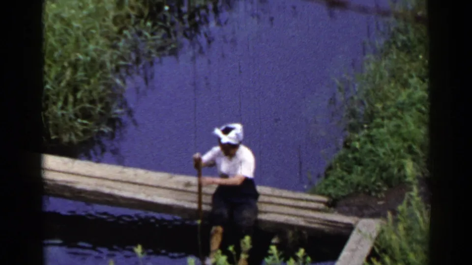 1953 Japan frame showing an adult man seated on a wooden bridge, holding a pole beside still water and green plants.