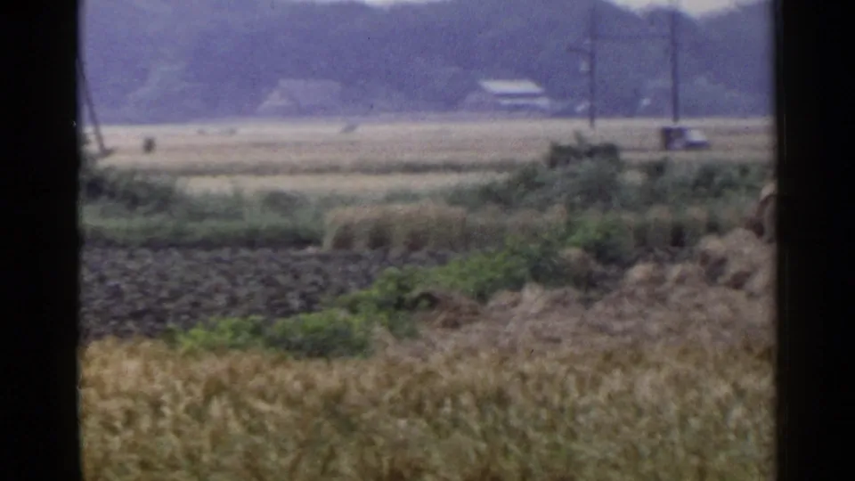 1953 Japan rural frame with dark plowed ground, green field patches, utility poles, a small distant vehicle, and a hazy tree line.