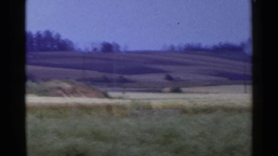 1953 Japan 8mm frame of a rural field, brush pile, low hillside, and dark tree line under a blue-purple sky.