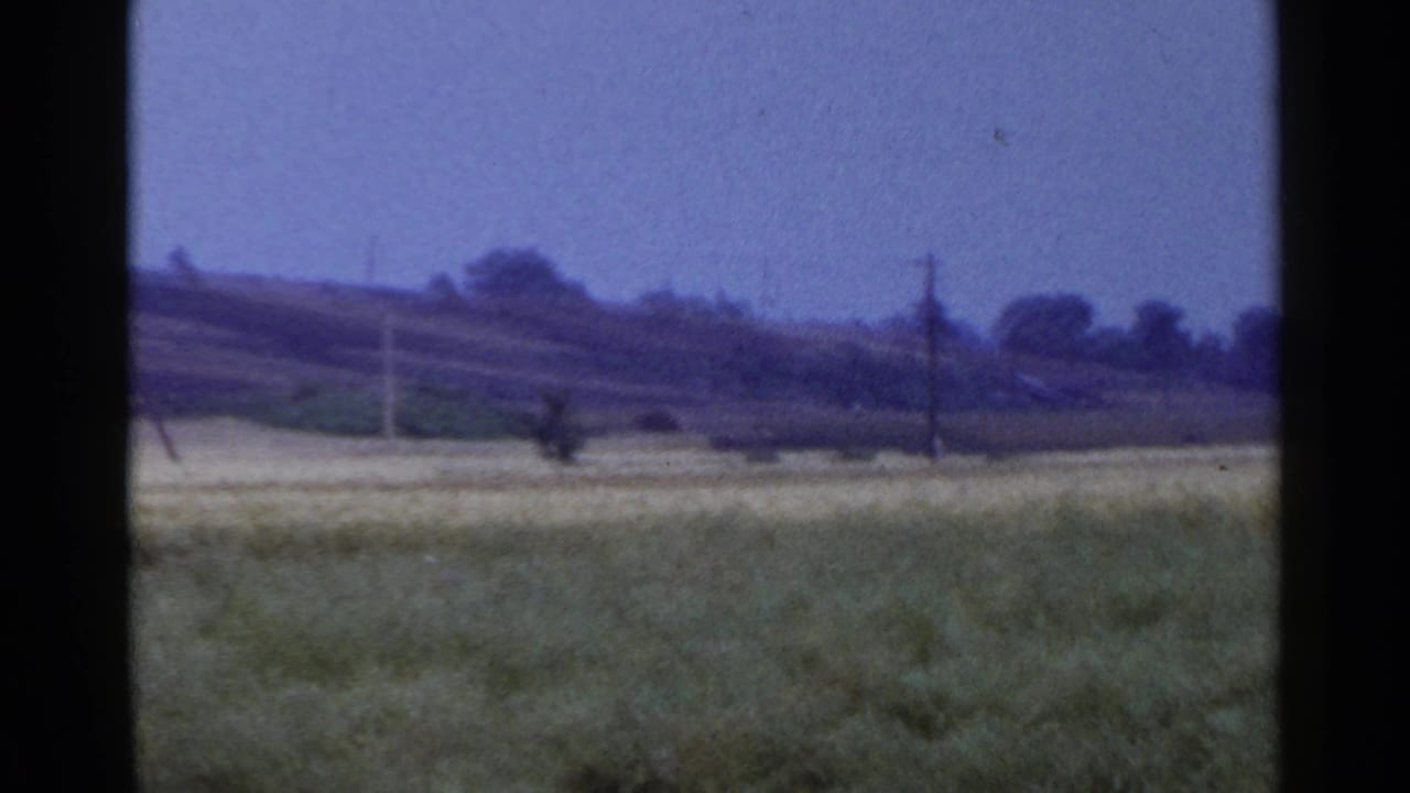 1953 8mm rural view of an open field, distant utility poles, trees, and low hills seen through dark film-frame edges.