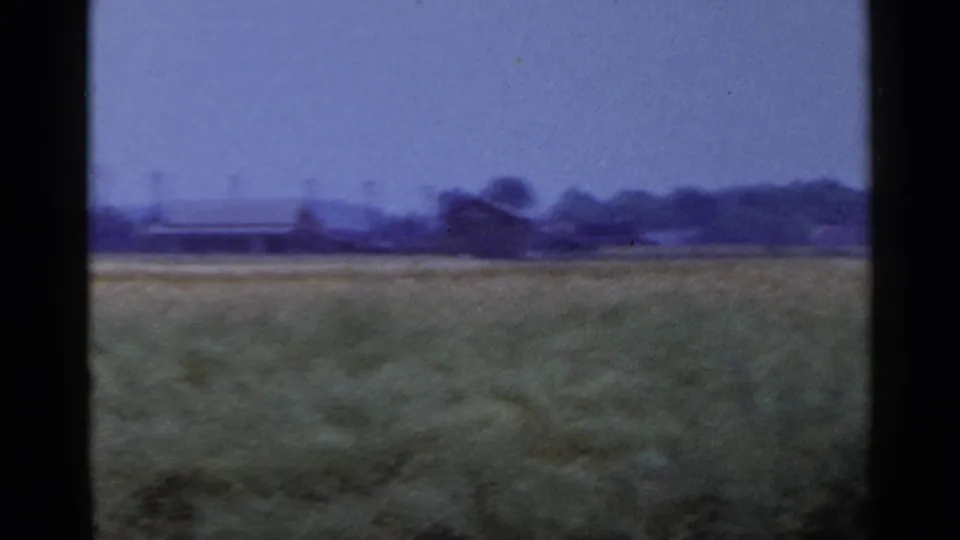 1953 home-movie frame of an open rural field with a long low structure, distant trees, and soft 8mm color shift.