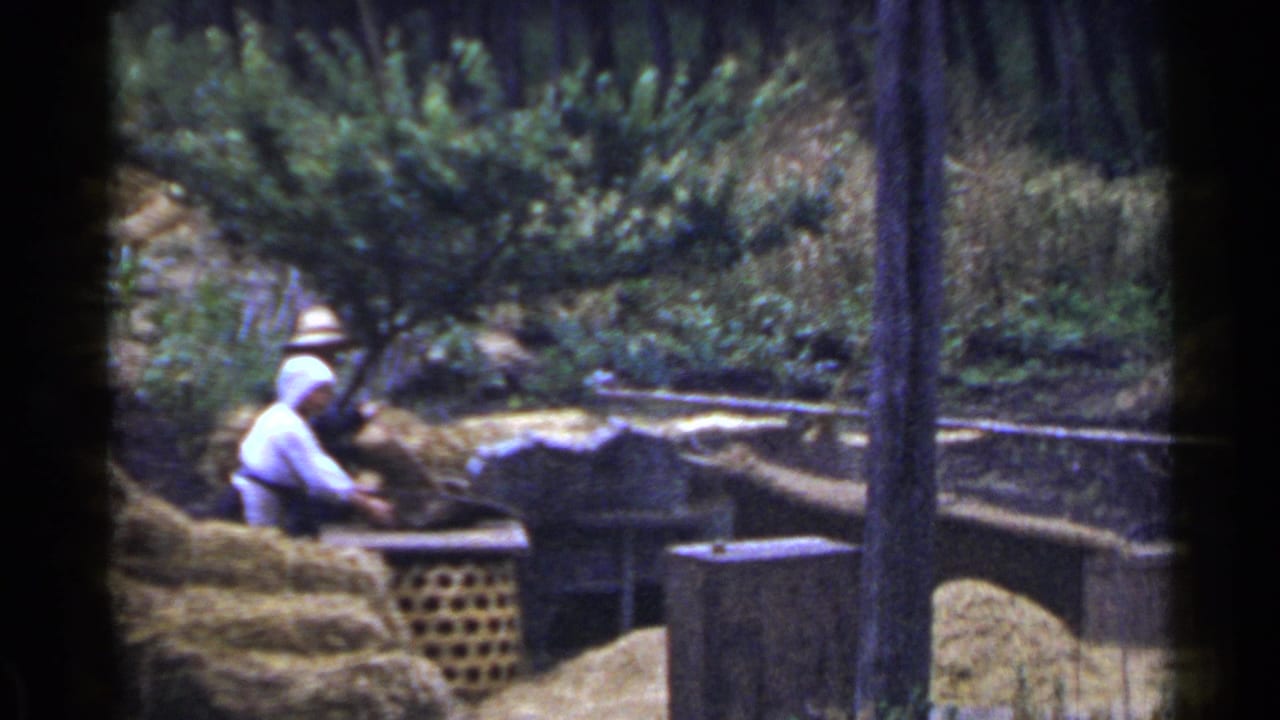 Two adults work near straw piles and wooden structures in a leafy outdoor setting.