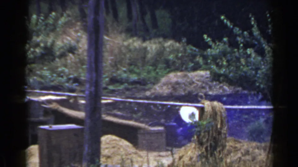 1953 Japan home-movie frame with a white-covered worker bending beside a tall straw or hay pile and wooden rails.