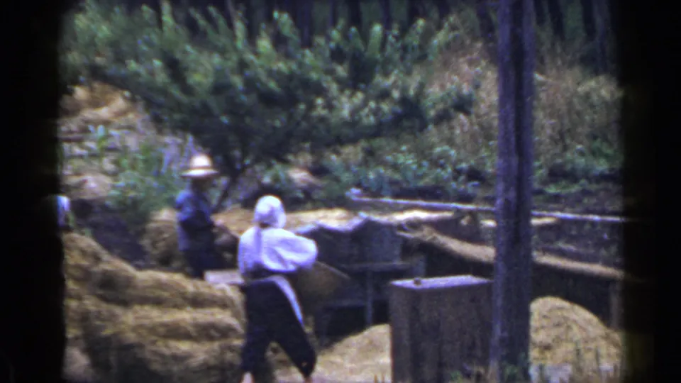 1953 Japan 8mm frame showing adults near grain or hay piles, with one person in a white bonnet and apron and another in a hat.