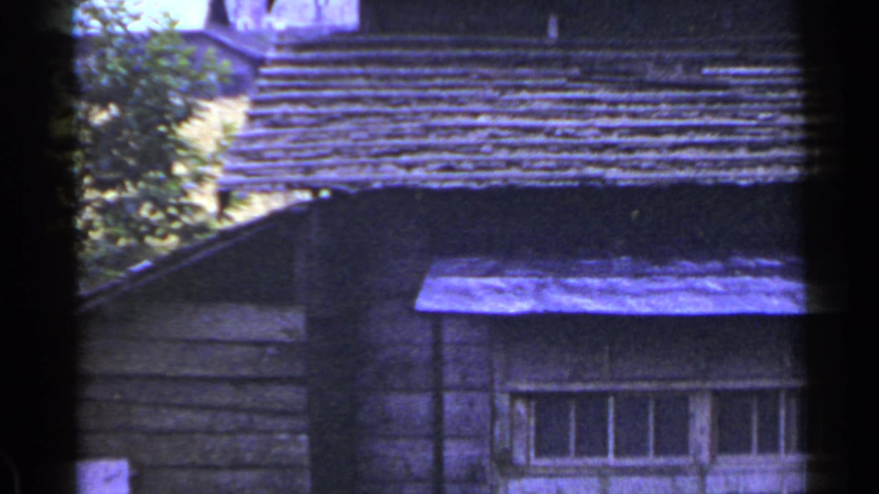 Upper portion of an old wooden building with a long window, roofline, and trees behind it.