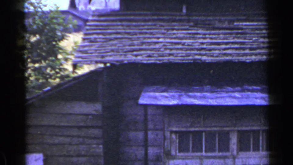 1953 Japan home-movie frame with an upper wooden wall, long multi-pane window, sloped roofline, and trees in the background.