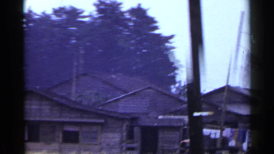 1953 Japan 8mm frame showing wooden rural buildings, tall utility poles, and a tree line beyond the farm landscape.