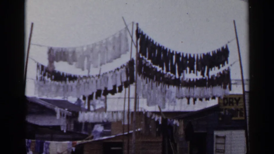 1953 Japan residential frame with hanging garments, clothesline poles, rooftops, and a partial unreadable sign at right.