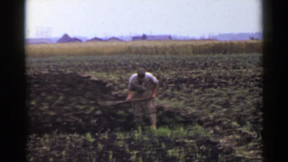 The 1953 Japan home-movie frame shows a farmer pulling a hoe through dark rows, with open farmland and low structures in the background.