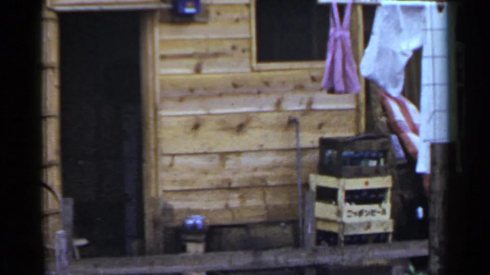 1953 Japan home-movie frame showing laundry hanging beside a wooden cabin entrance with crates and household objects nearby.