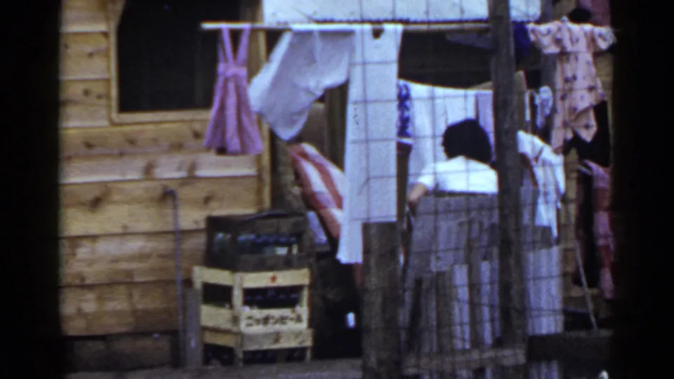 1953 Japan home-movie frame with a woman working near laundry outside a wooden cabin, with clotheslines, crates, and posts visible.