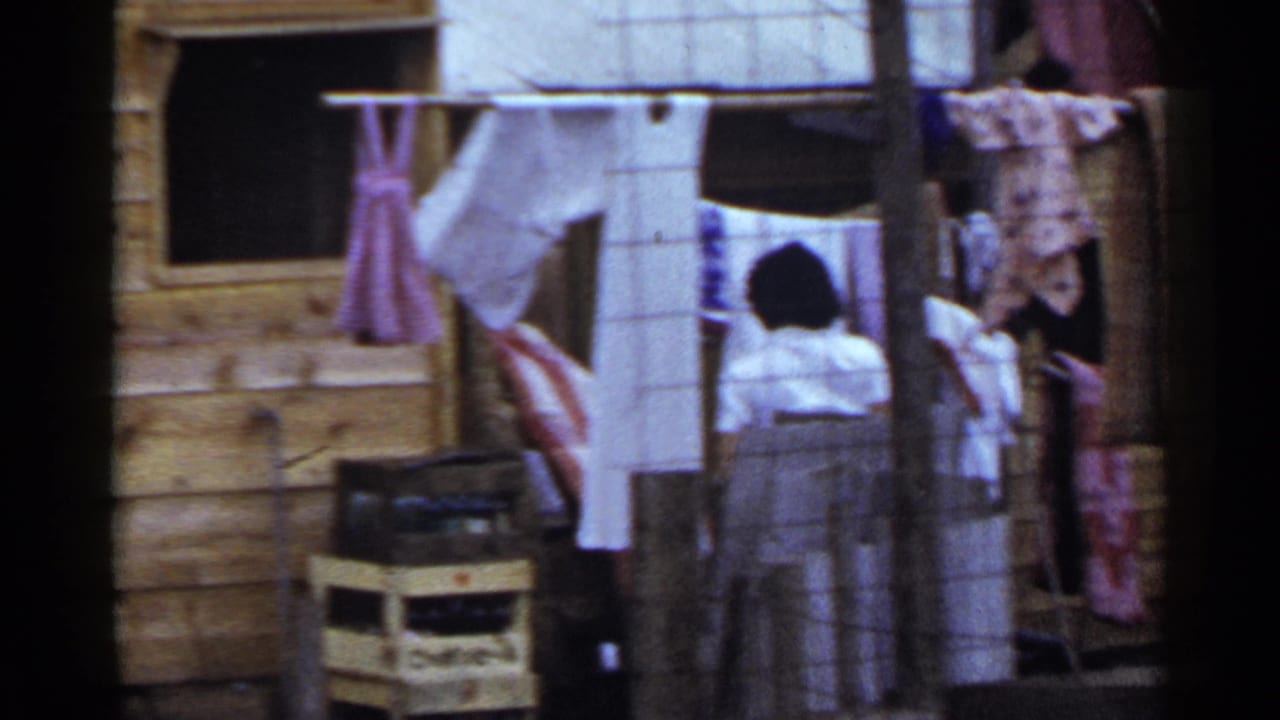 1953 Japan home-movie frame showing a woman by hanging laundry outside a wooden house, with crates, posts, and garments in view.
