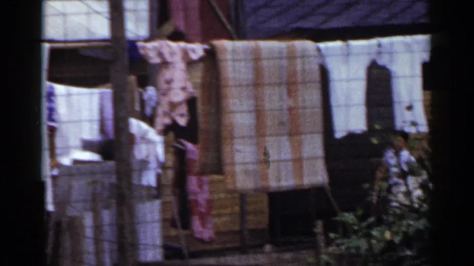 1953 Japan home-movie frame showing laundry on lines, wooden exterior walls, posts, and a small figure partly hidden near plants.