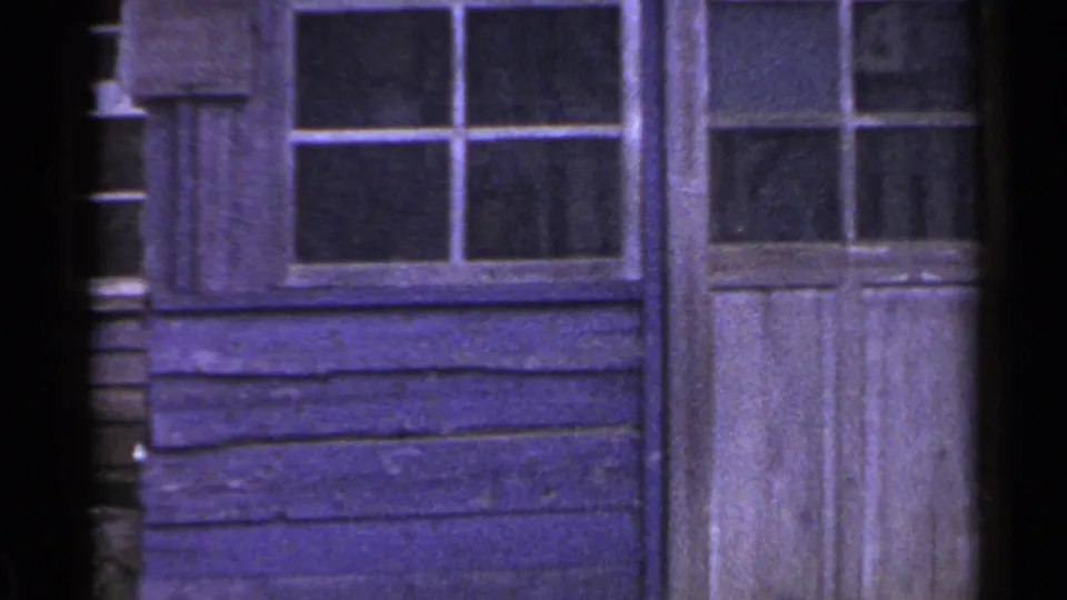 1953 Japan home-movie frame showing a purple-toned wooden wall, small-paned windows, and a close view of a weathered doorway.