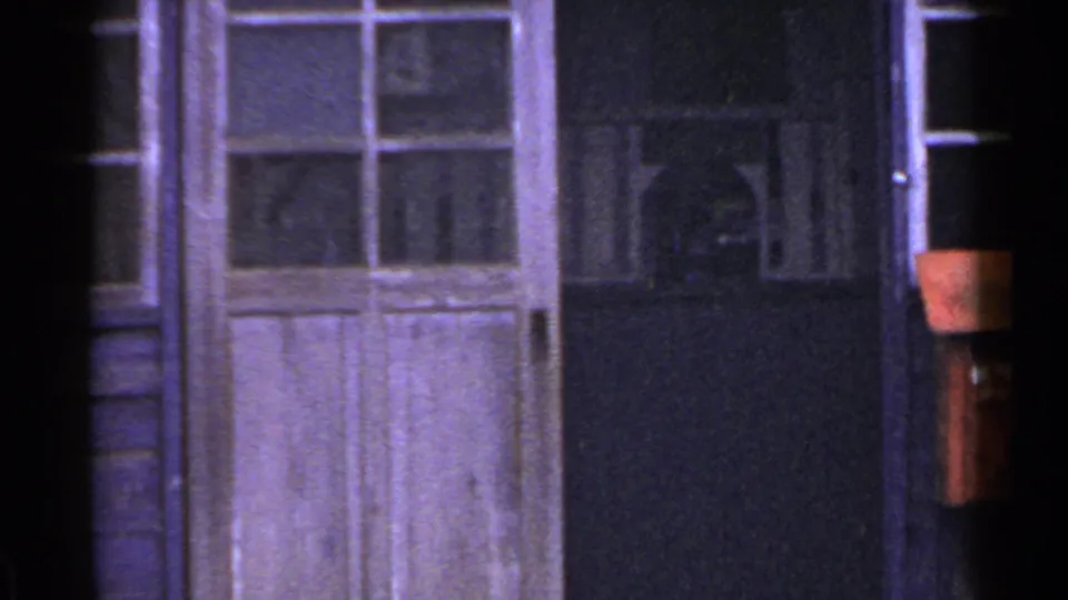 1953 Japan home-movie frame showing a close exterior view of a wooden door, window, dark entry area, and edge of a red mailbox.
