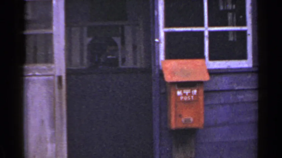 1953 Japan home-movie frame showing a red mailbox with Japanese characters mounted on a weathered wooden exterior beside a window and doorway.